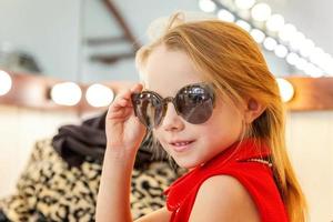 pequeño niña en Gafas de sol cerca espejo foto