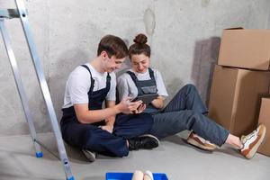 House DIY renovation concept. Young happy couple sitting on floor looking home repair plan in tablet indoors. Woman showing to man plan interior design online. Couple in new home during repair works. photo