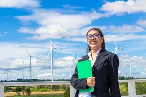 Energy engineer female working at wind farm. asian business woman wearing suit holding tablet and clipboard with windmill environment renewable energy. photo