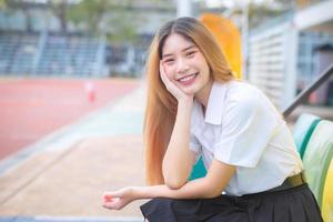 retrato de linda asiático tailandés joven niña estudiante mirando simpático es sentado sonriente felizmente confidente y mano participación cara como bueno a presente alguna cosa instalaciones estadio en Universidad antecedentes. foto