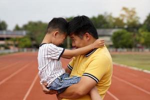 The father held his son by the waist, both of them facing each other, their foreheads facing each other, laughing. happy smile Evening time in the stadium, in the concept of family activities. photo