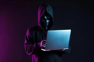 a hacker in a hoodie using a laptop computer isolated on dark background with neon light. photo