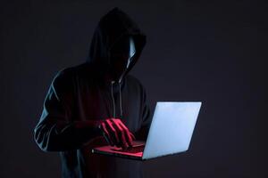 a hacker in a hoodie using a laptop computer isolated on dark background with neon light. photo