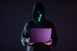 a hacker in a hoodie using a laptop computer isolated on dark background with neon light. photo