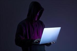 a hacker in a hoodie using a laptop computer isolated on dark background with neon light. photo