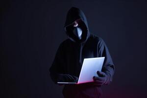 a hacker in a hoodie using a laptop computer isolated on dark background with neon light. photo