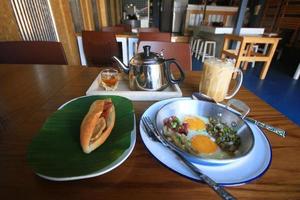 Indochina freído en sarten huevo con coberturas con junquillo un pan emparedado con queso, jamón en Fresco verde plátano hoja y hielo café, té caliente en de madera mesa en hecho en casa tailandés estilo foto