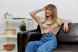 Cute smiling blonde lady in casual sweater looks aside enjoy good mood sitting in sofa at modern living room interior. Female client have shrink appointment in psychological help office. Copy space photo