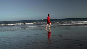 Happy young woman in red dress runs along the ocean beach at sunset. Concept of carefree modern life. Slow motion video