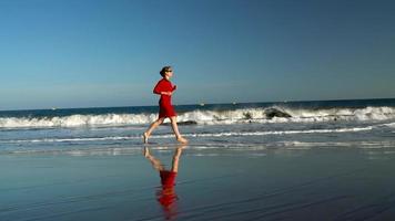 content Jeune femme dans rouge robe court le long de le océan plage à le coucher du soleil. concept de insouciant moderne vie. lent mouvement video