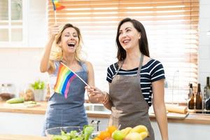 Happy caucasian LGBT couple, Lesbian holding and waving rainbow LGBT Pride flag together in the kitchen at home. Diversity of LGBT relationships. Cheerful Homosexual couple. photo