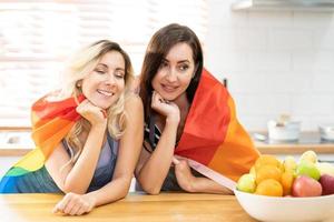 Happy caucasian LGBT couple, Lesbian holding and waving rainbow LGBT Pride flag together in the kitchen at home. Diversity of LGBT relationships. Cheerful Homosexual couple. photo