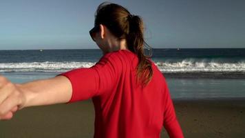 seguir yo - contento joven mujer en rojo vestir tracción tipo mano. mano en mano caminando a el agua en el Oceano playa a video