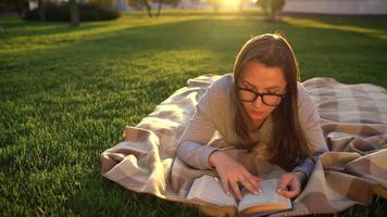 Girl in glasses reading book lying down on a blanket in the park at sunset video