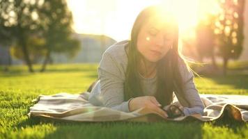 menina dentro óculos lendo livro deitado baixa em uma cobertor dentro a parque às pôr do sol video