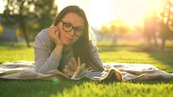 Girl in glasses reading book lying down on a blanket in the park at sunset video