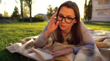 fille dans des lunettes en train de lire livre mensonge vers le bas sur une couverture dans le parc à le coucher du soleil video