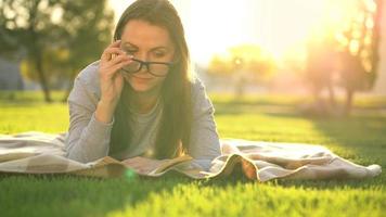 menina dentro óculos lendo livro deitado baixa em uma cobertor dentro a parque às pôr do sol video