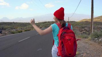 viajero mujer autoestop en un soleado la carretera y caminando. mochilero mujer mirando para un paseo a comienzo un viaje en un iluminado por el sol país la carretera. lento movimiento video