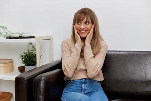 Joyful funny blonde lady in casual sweater touches cheeks looks aside sitting in sofa at modern living room interior. Female client have shrink appointment in psychological help office. Copy space photo