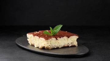 A piece of tiramisu sprinkled with cocoa, on top of a sprig of fresh mint on a black background photo