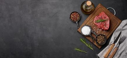 Raw piece of beef with spices pepper, rosemary sprig, salt and olive oil on a wooden board, black background photo