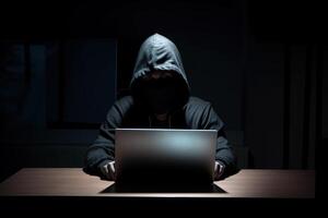 Hacker wearing black hoodie using laptop computer while working on dark background. photo