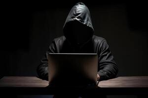 Hacker wearing black hoodie using laptop computer while working on dark background. photo