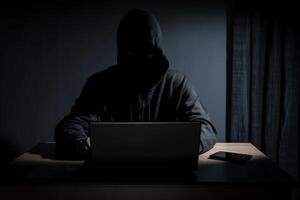 Hacker wearing black hoodie using laptop computer while working on dark background. photo
