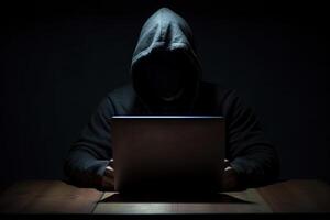 Hacker wearing black hoodie using laptop computer while working on dark background. photo