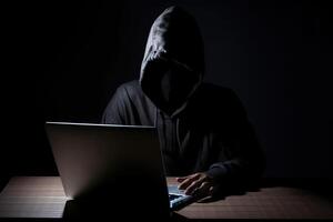 hacker vistiendo negro capucha utilizando ordenador portátil computadora mientras trabajando en oscuro antecedentes. ai generado foto