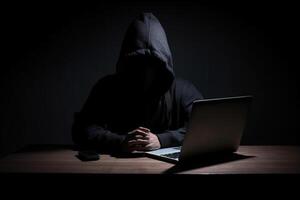 Hacker wearing black hoodie using laptop computer while working on dark background. photo