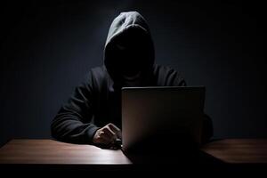 Hacker wearing black hoodie using laptop computer while working on dark background. photo