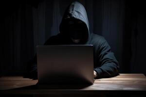 Hacker wearing black hoodie using laptop computer while working on dark background. photo