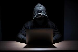 Hacker wearing black hoodie using laptop computer while working on dark background. photo