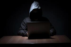 Hacker wearing black hoodie using laptop computer while working on dark background. photo