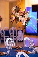 Festive table with blue cloth, napkins, plates, cutlery and glasses prepared for wedding guests in restaurant. Beautiful floral arrangement in gold metal vase with number four. Amazing servings photo