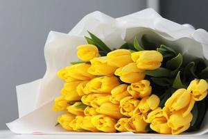 beautiful big bouquet of yellow tulips in a white package on white table and gray wall background close up. Congratulation. Festive background. copy space. photo
