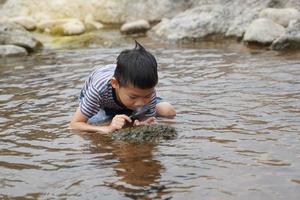 Asian boy enjoys exploring creatures in stream, along rocks with magnifying glass. concept of learning outside the classroom, natural learning resources. Soft and selective focus. photo