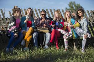 A group of people dressed in the style of the nineties sit on a bench by a skewed fence. photo
