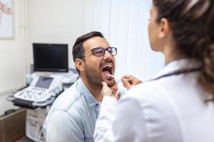 A young man sits on an exam table across from his doctor. The doctor reaches forward with a tongue depressor as the man looks up and sticks out his tongue. photo