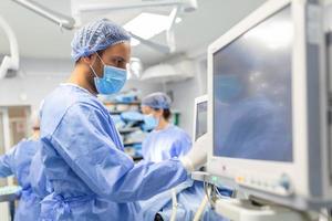 Anesthetist Working In Operating Theatre Wearing Protecive Gear checking monitors while sedating patient before surgical procedure in hospital photo