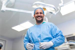 Portrait of male nurse surgeon OR staff member dressed in surgical scrubs gown mask and hair net in hospital operating room theater making eye contact smiling pleased happy looking at camera photo