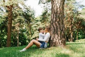 joven caucásico mamá y su tres años hijo tener divertido sentado en el parque cerca un árbol en el césped en verano foto