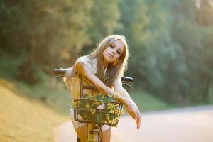 A girl in white shorts and a shirt with a bicycle and a basket of flowers stands in the park in the evening against the background of the setting sun photo