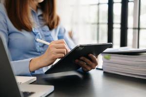 mujer de negocios que trabaja con una tableta de pantalla ancha en blanco vr, una computadora y un teléfono inteligente en el fondo borroso de la oficina. foto