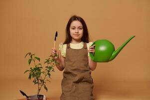 Adorable smiling little kid girl holding watering can and garden shovel, learns gardening, isolated on beige background photo