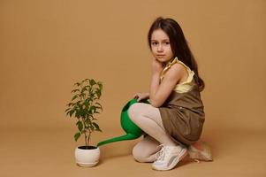 Conscious 6 years old kid girl takes care of flowering seedling, watering it using a watering can over beige background photo