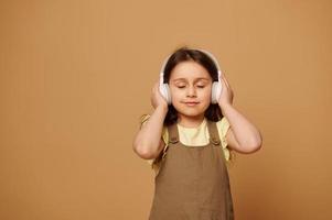 contento pequeño niña escucha a banda sonora en auriculares, disfruta calmante música con su ojos cerrado terminado beige antecedentes foto