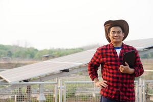 Asian man farmer wears hat, red plaid shirt, holds smart tablet, stands beside solar panel. Concept, farmer installed photovoltaic for use in agriculture for electricity.  Smart farmer. photo